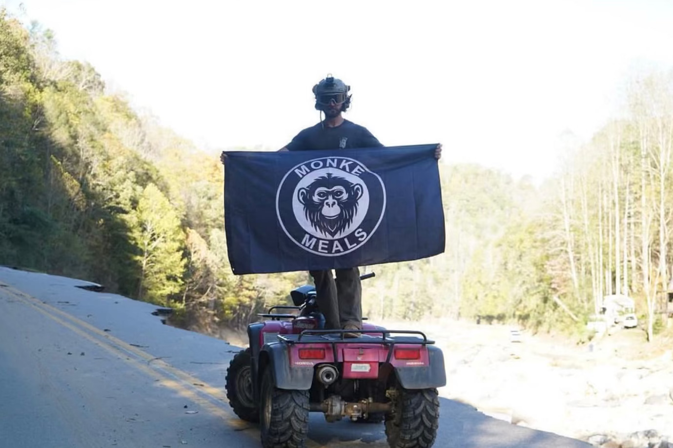 Person on an ATV holding a flag with 'Monk Meals' logo in a forest setting